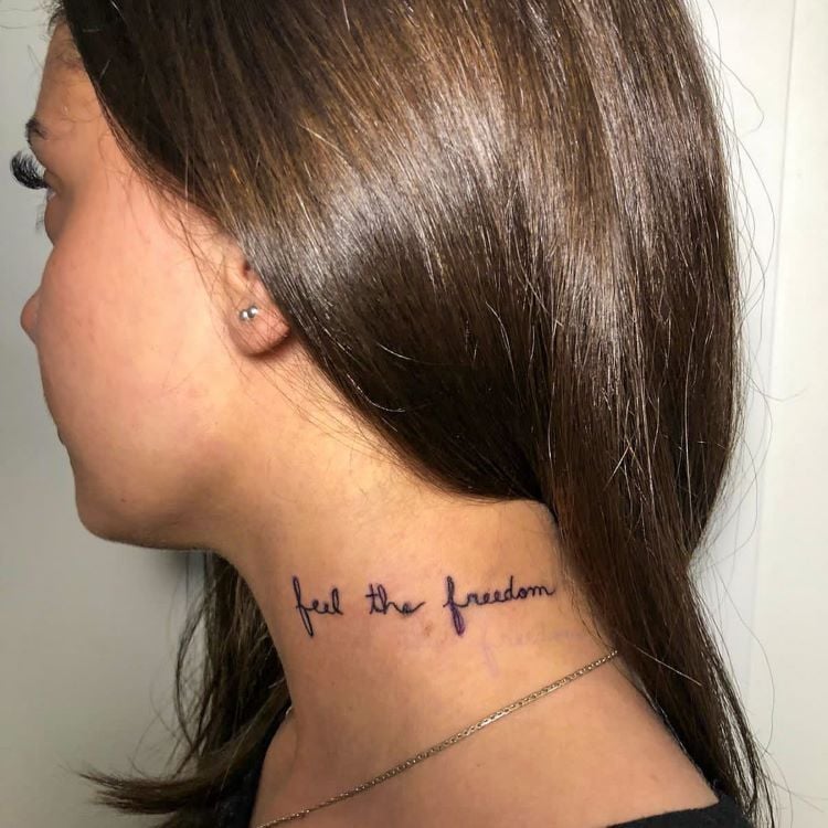 A imagem mostra uma pessoa de perfil com cabelo liso e castanho. Ela tem uma tatuagem no pescoço com a frase "feel the freedom" escrita em letras cursivas pretas. A tatuagem está localizada na lateral do pescoço, logo abaixo da linha do cabelo, e a pessoa usa um brinco pequeno.