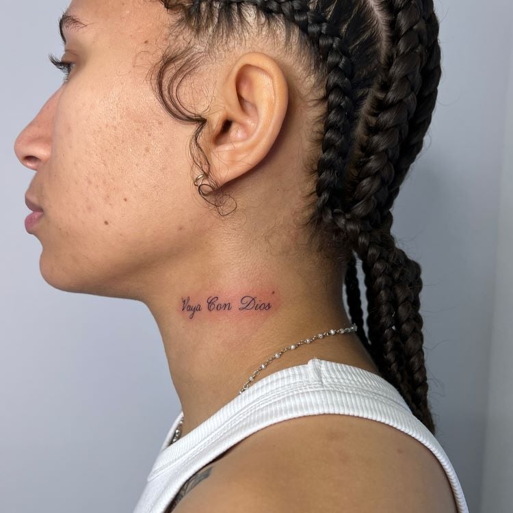 A imagem mostra uma pessoa de perfil com cabelo trançado. Ela tem uma tatuagem no pescoço com a frase "Vaya Con Dios" escrita em letras cursivas pretas. A tatuagem está localizada na lateral do pescoço, logo abaixo da linha do maxilar. A pessoa usa uma corrente prateada.