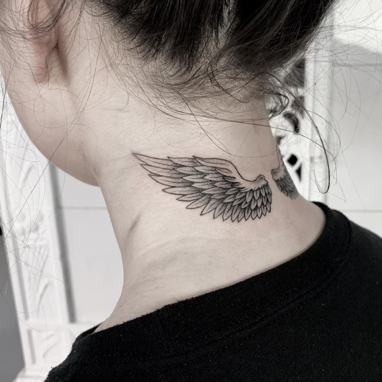 A imagem mostra uma pessoa de costas com cabelo preso. Ela tem uma tatuagem no pescoço de asas de anjo estendendo-se horizontalmente, detalhadas em preto. A tatuagem está localizada na base do pescoço, logo abaixo da linha do cabelo, e é feita com traços finos e delicados.