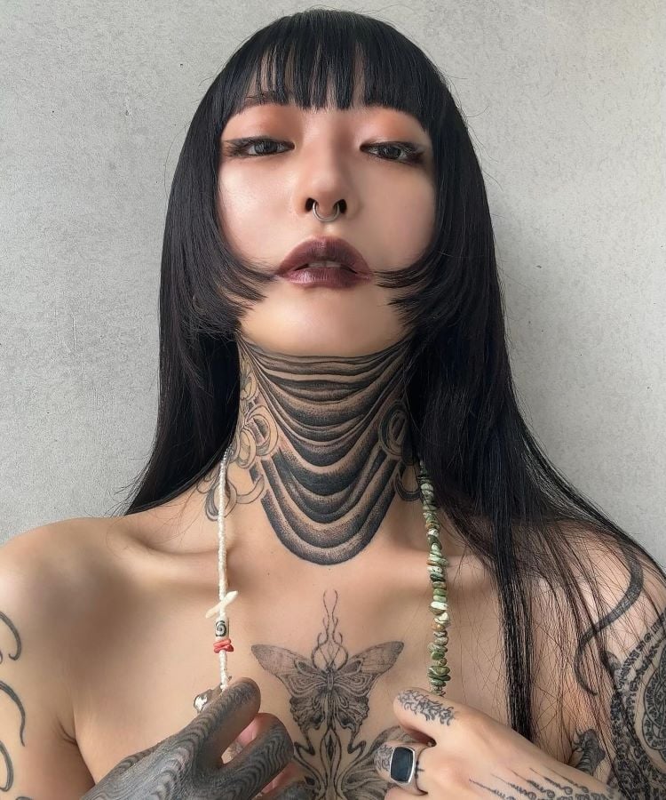 A imagem mostra uma pessoa de frente com cabelo preto liso e franja. Ela tem uma tatuagem no pescoço com um design de várias faixas pretas curvas. A pessoa usa maquiagem marcante, um piercing no septo e segura um colar de contas brancas.