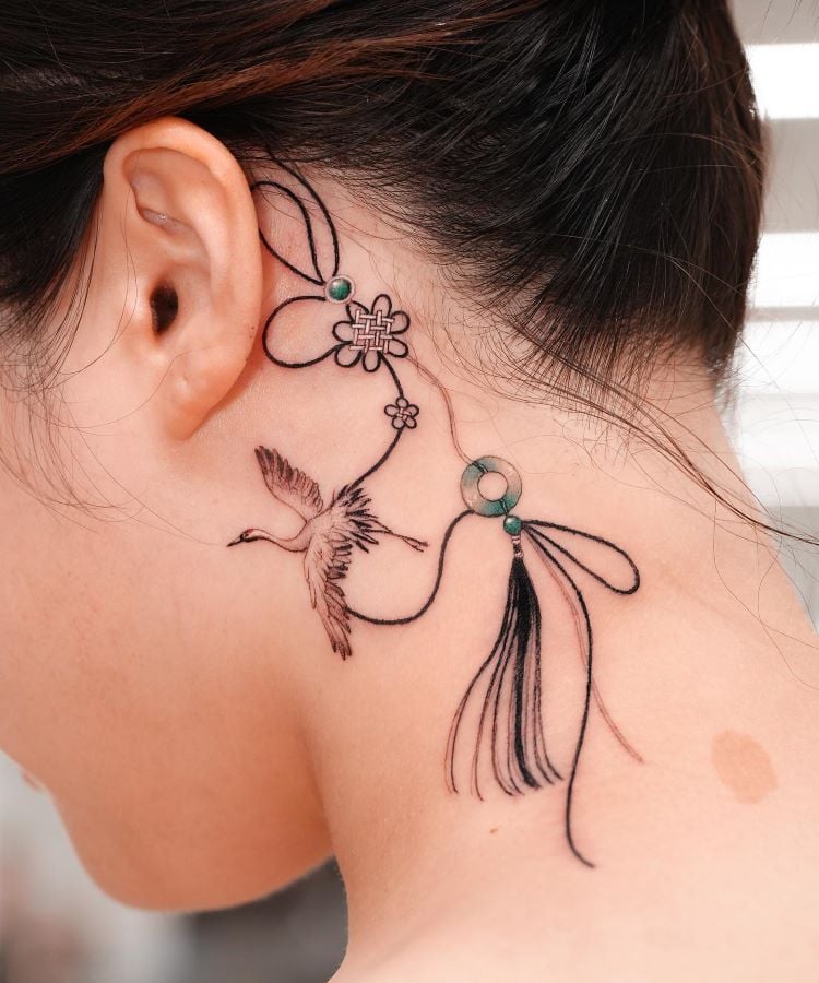 A imagem mostra uma pessoa de perfil com cabelo preso. Ela tem uma tatuagem no pescoço de um pássaro voando, flores e linhas curvas, detalhadas em preto com pequenos pontos verdes. A tatuagem começa atrás da orelha e se estende pela lateral do pescoço.