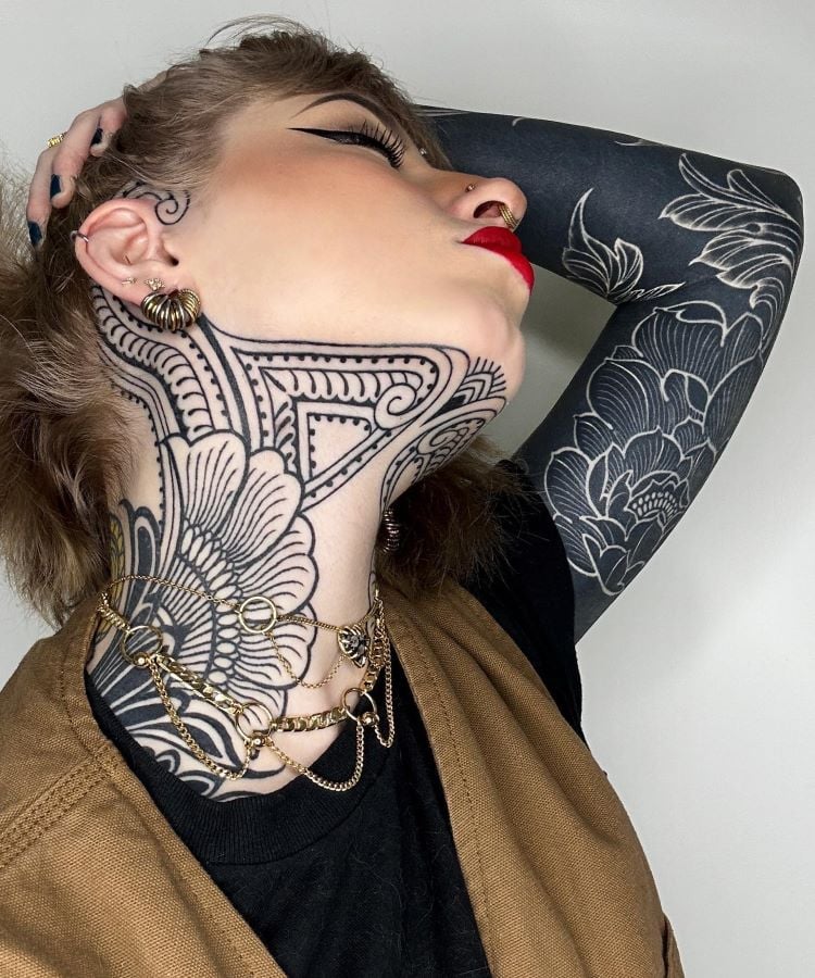 A imagem mostra uma pessoa com a cabeça inclinada para trás, exibindo uma tatuagem no pescoço com desenhos detalhados em preto. Ela tem maquiagem marcante, batom vermelho e piercings. O braço direito também é tatuado com flores. Usa brincos dourados e colares de corrente.