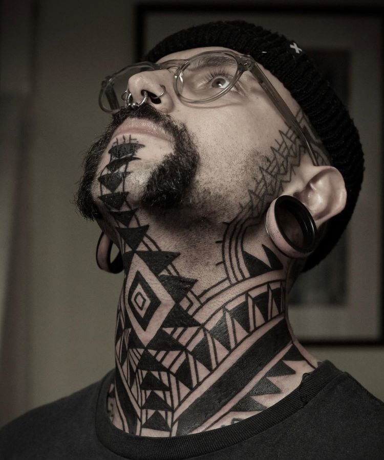 A imagem mostra uma pessoa com óculos, barba e alargadores nas orelhas. Ela tem uma tatuagem no pescoço de padrões geométricos em preto, que cobrem desde o queixo até a clavícula. As formas incluem triângulos, losangos e linhas, criando um design tribal intrincado e simétrico.