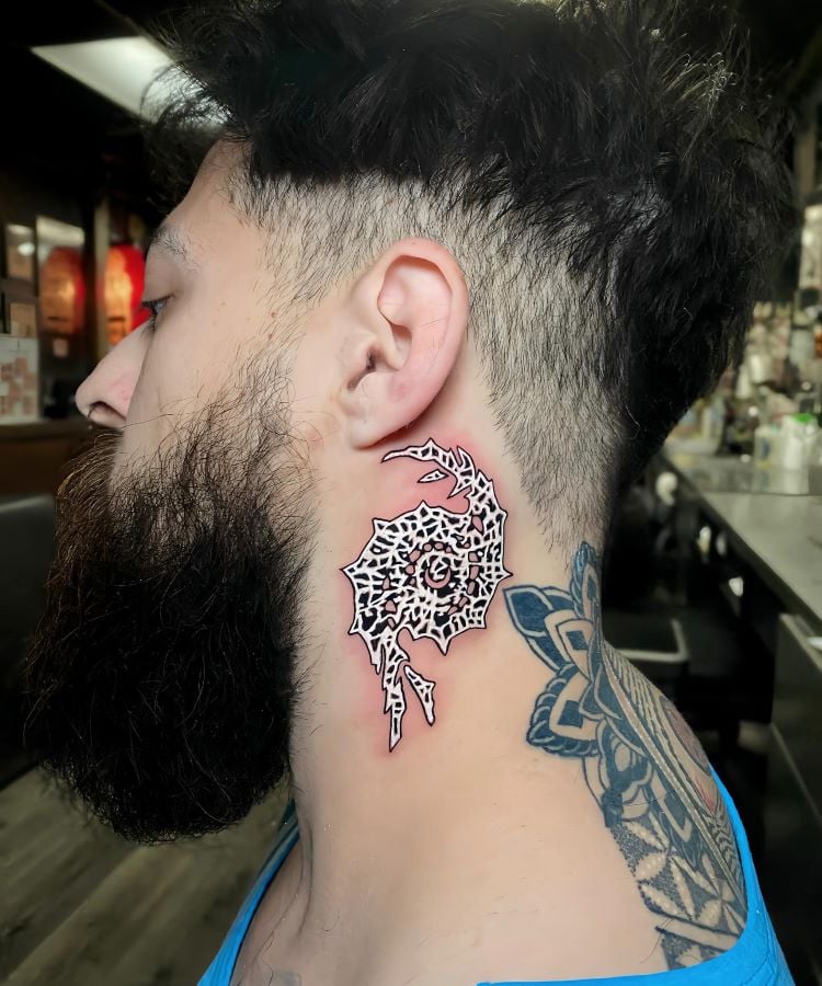 A imagem mostra uma pessoa de perfil com cabelo curto e barba. Ela tem uma tatuagem no pescoço de um design abstrato em preto e vermelho, parecendo um padrão tribal. A pessoa também tem uma tatuagem geométrica no ombro, visível pela camiseta azul cavada.