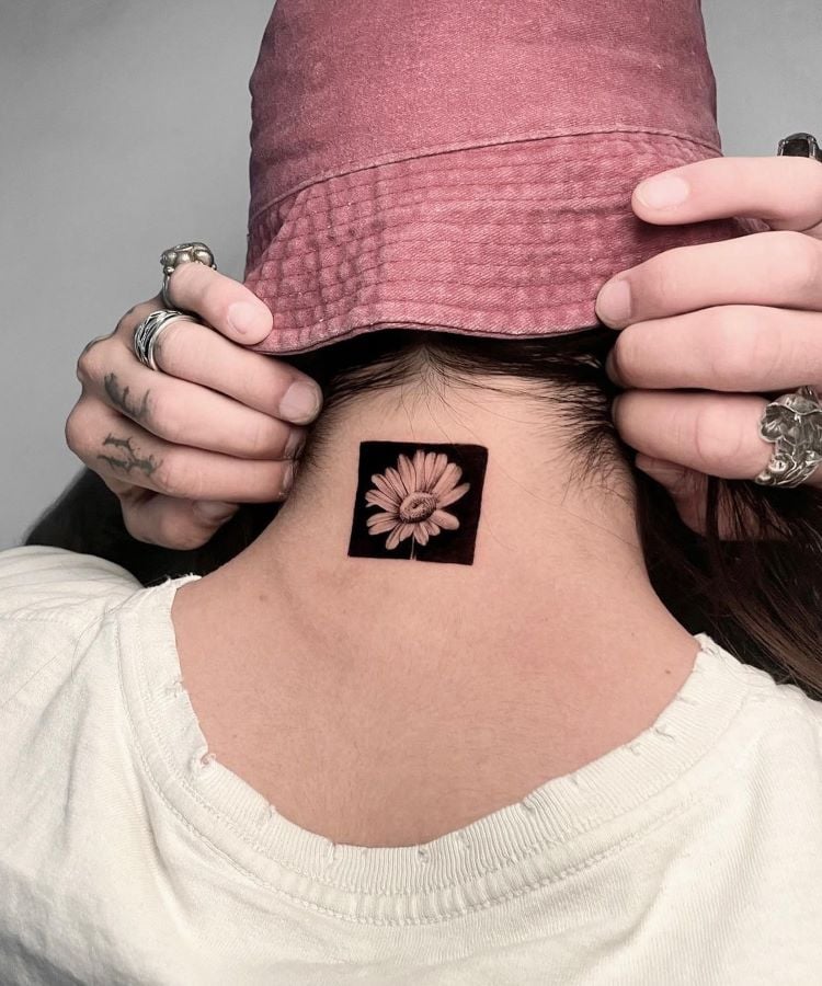 A imagem mostra uma pessoa de costas usando um chapéu rosa. Ela tem uma tatuagem no pescoço de uma flor detalhada dentro de um quadrado preto. A pessoa está segurando o chapéu com ambas as mãos, revelando vários anéis nos dedos. A tatuagem é centralizada na nuca.