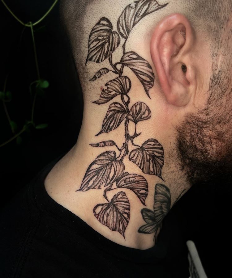 A imagem mostra uma pessoa de perfil com uma tatuagem no pescoço de uma planta trepadeira com folhas detalhadas em preto. A pessoa tem barba e usa uma camiseta preta. A tatuagem se estende da base do pescoço até atrás da orelha.