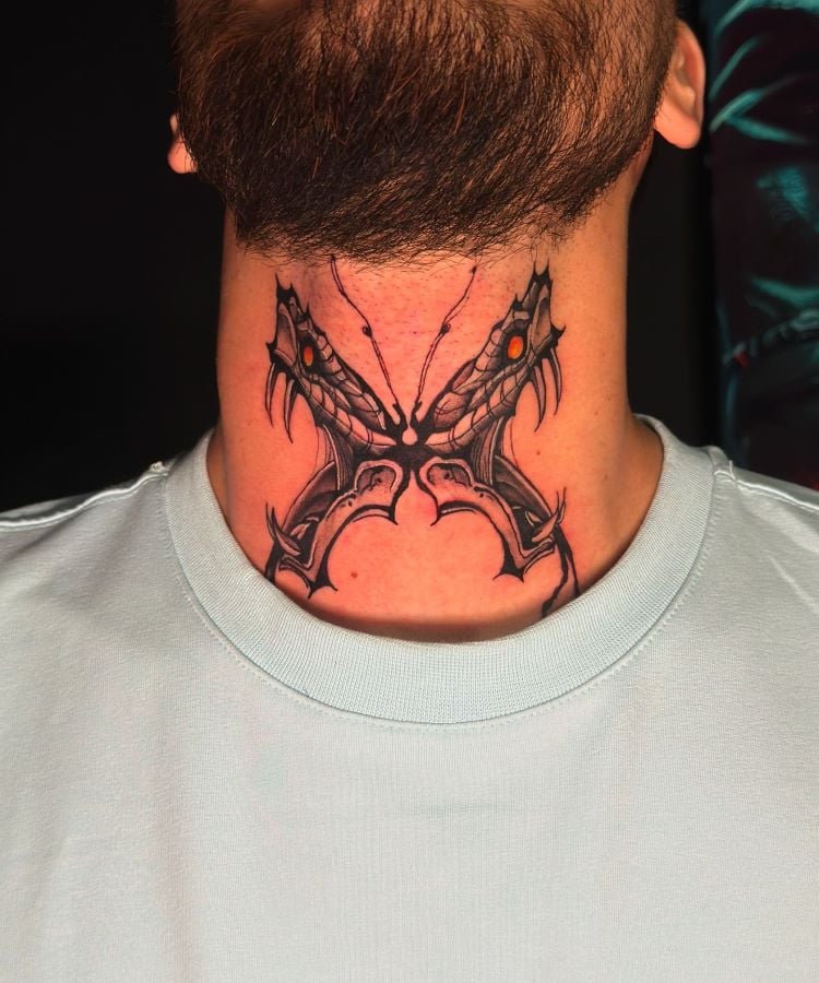 A imagem mostra uma pessoa com barba e camiseta clara. Ela tem uma tatuagem no pescoço de um design simétrico e abstrato em preto, com detalhes em vermelho. A tatuagem parece ter duas cabeças de dragão opostas, com garras e olhos brilhantes, cobrindo a parte frontal do pescoço.