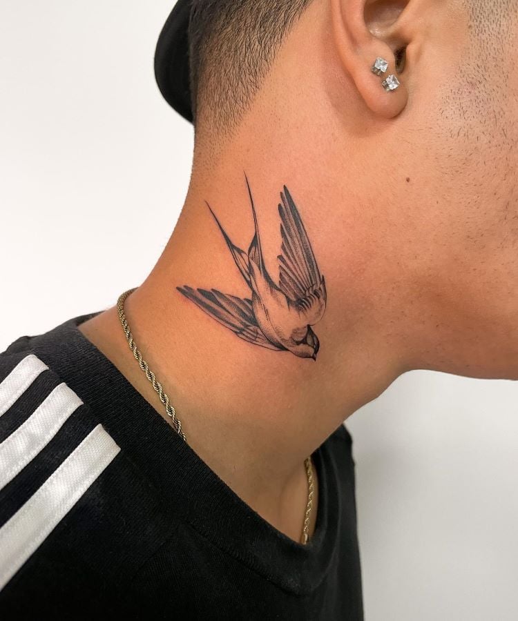 A imagem mostra uma pessoa de perfil com cabelo curto e uma corrente dourada. Ela tem uma tatuagem no pescoço de uma andorinha em voo, desenhada em preto e cinza, com detalhes realistas nas asas e no corpo. A pessoa usa brincos brilhantes e uma camiseta preta com listras brancas.