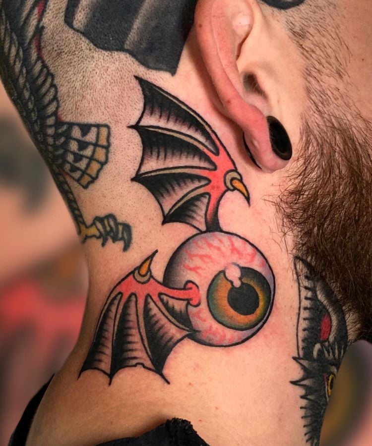 A imagem mostra uma pessoa de perfil com barba e alargador preto na orelha. Ela tem uma tatuagem no pescoço de um olho com asas de morcego em estilo de desenho animado, com cores vivas como vermelho, preto e amarelo. A tatuagem está localizada na lateral do pescoço.