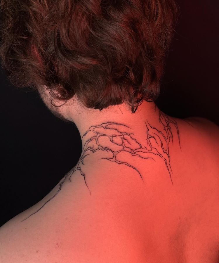 A imagem mostra uma pessoa de costas com cabelo castanho ondulado. Ela tem uma tatuagem no pescoço de galhos finos e detalhados, desenhados em linhas pretas que se estendem da base do crânio até os ombros. A tatuagem é delicada e parece parte de uma árvore.