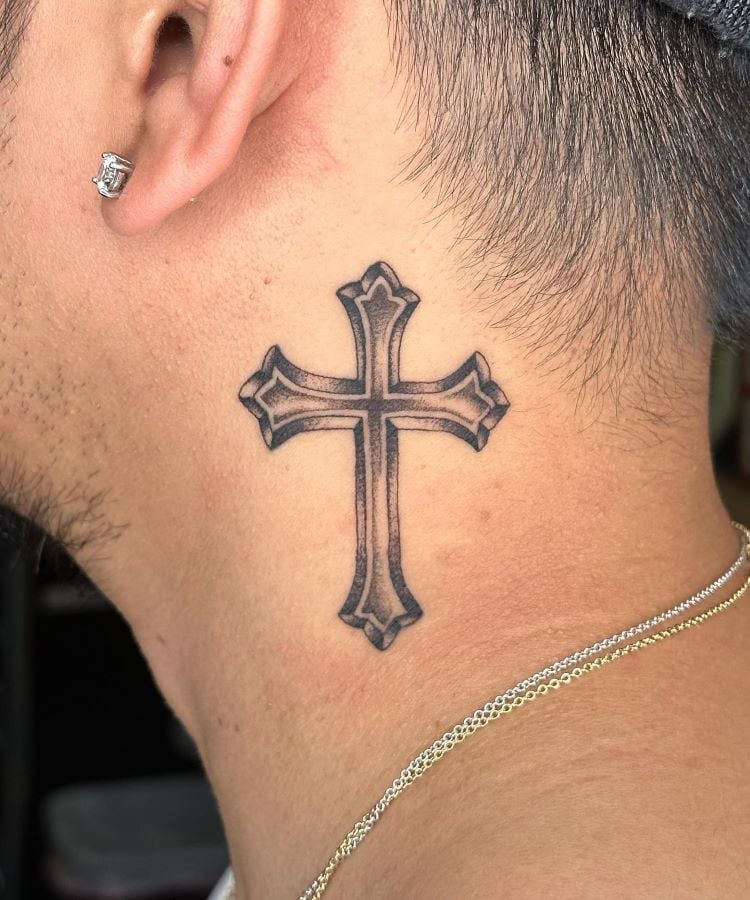 A imagem mostra uma pessoa de perfil usando um brinco pequeno e brilhante. Ela tem uma tatuagem no pescoço de uma cruz detalhada em preto, localizada logo abaixo da orelha. A pessoa também usa um colar fino de corrente dourada.