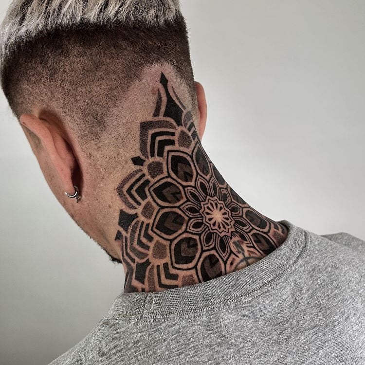 A imagem mostra uma pessoa de costas com cabelo curto raspado nas laterais. Ela tem uma tatuagem no pescoço de um mandala geométrico detalhado em preto, que se estende da base do crânio até a parte superior das costas. A tatuagem é simétrica e intricada.