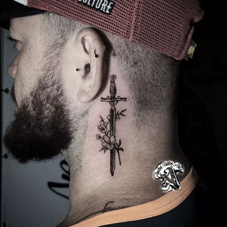 A imagem mostra uma pessoa de perfil usando um boné vermelho e preto. Ela tem uma tatuagem no pescoço de uma espada vertical cercada por flores. A pessoa tem barba e usa vários piercings na orelha.