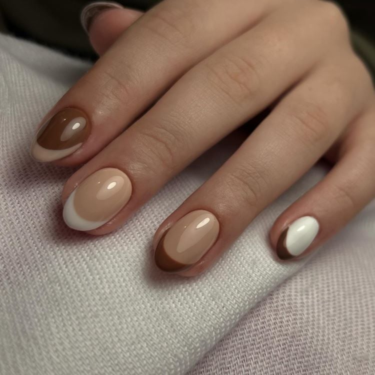 A imagem mostra uma mão com unhas pintadas em um design de manicure francesa invertida. A cor predominante nas unhas é marrom, com a ponta em branco contrastante. 