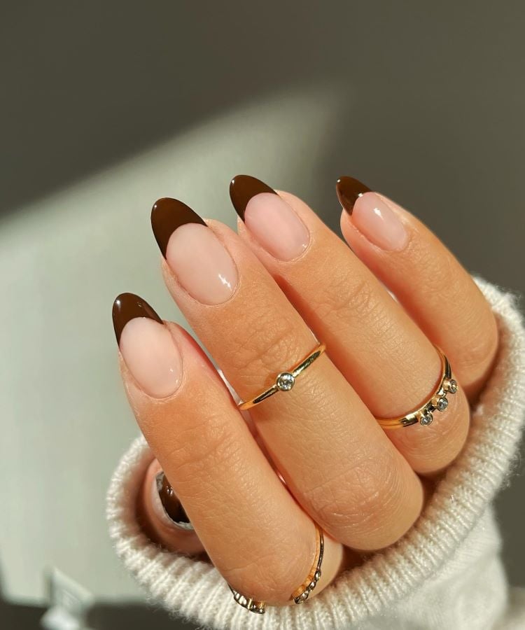 A imagem mostra uma mão com unhas pintadas em um design de manicure francês, onde a ponta é de cor marrom escura. As unhas têm formato amendoado e são longas. A mão exibe 3 anéis delicados, um no dedo anelar, outro no dedo médio e outro no outro, os 3 com pequenas pedras brilhantes.