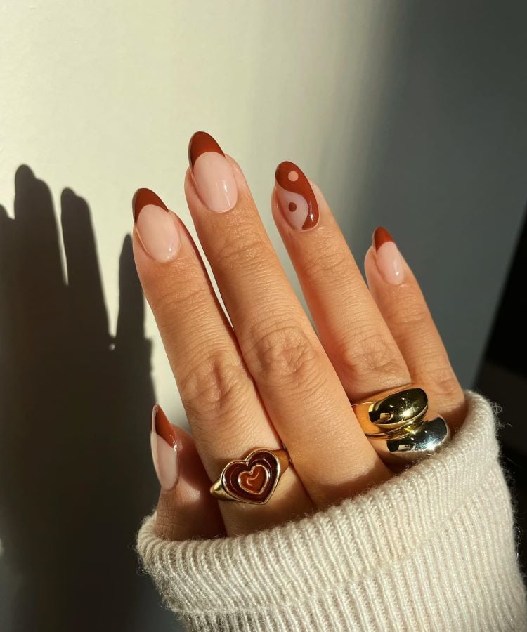 A imagem mostra uma mão com unhas pintadas em um design de manicure francês, com a ponta em marrom e detalhes adicionais na mesma cor. A mão está adornada com três anéis dourados, um dos quais tem um coração. A sombra da mão projeta-se sobre uma superfície clara.