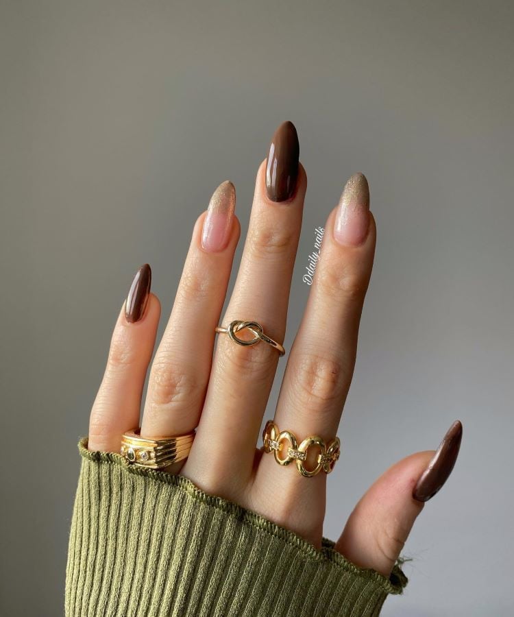 A imagem mostra uma mão com unhas pintadas em tons de marrom e nude, com a unha do dedo anelar destacando-se por ter um esmalte brilhante dourado. A mão exibe anéis dourados em três dedos, e o pulso está coberto pela manga verde de uma malha canelada.