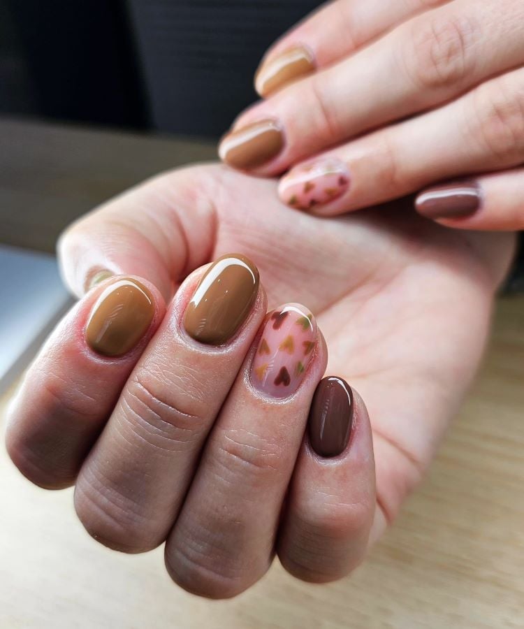 A imagem mostra uma mão com unhas pintadas em tons de marrom e uma unha decorada com um padrão que lembra corações, combinando tons de marrom e outros terrosos. A unha marrom no dedo anelar destaca-se com o design artístico, sugerindo uma tendência de moda em manicure.