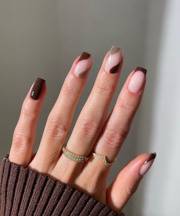 A imagem mostra uma mão com unhas pintadas em tons de marrom e nude, com uma unha marrom destacada no dedo anelar. As unhas têm formatos amendoado e uma delas possui detalhes brilhantes. A mão usa dois anéis dourados delicados e está sobre um fundo neutro.