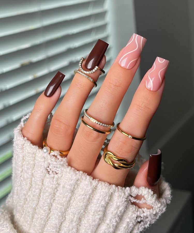 A imagem mostra uma mão com unhas postiças, algumas pintadas de marrom e outras com um design ondulado rosa e branco. A mão está adornada com vários anéis dourados, destacando-se contra um suéter branco felpudo.