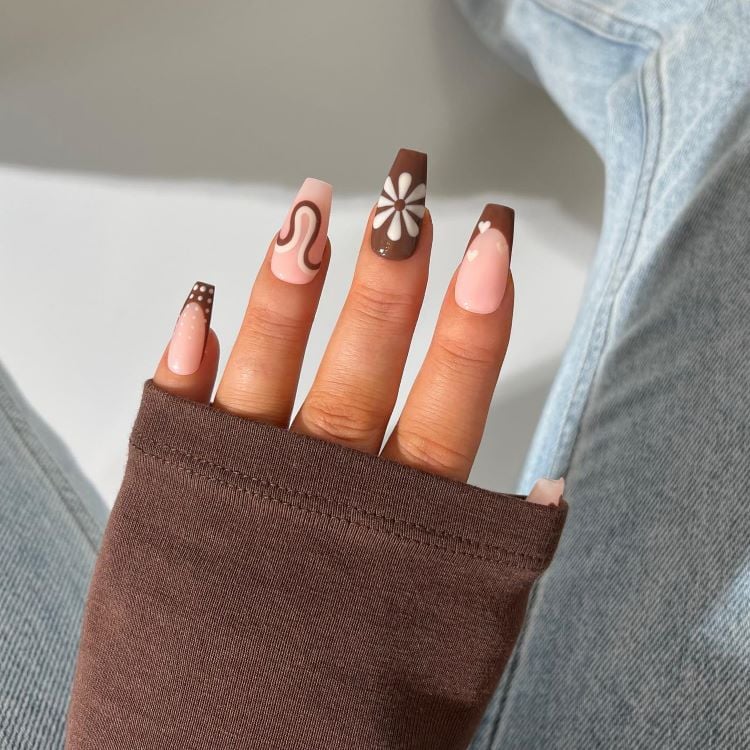 A imagem mostra uma mão com unhas compridas e bem cuidadas, apresentando um design artístico em tons de marrom e bege. A unha marrom combina com a roupa da pessoa, que parece ser uma manga de tecido grosso na cor marrom. A mão repousa sobre um tecido azul claro.