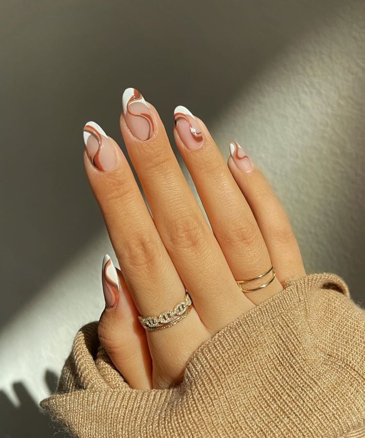 A imagem mostra uma mão com unhas pintadas com um design artístico que inclui linhas onduladas em tons de marrom, branco e rosa. A mão está adornada com anéis delicados e repousa sobre um tecido bege, sob uma luz suave que realça a unha marrom e o design.