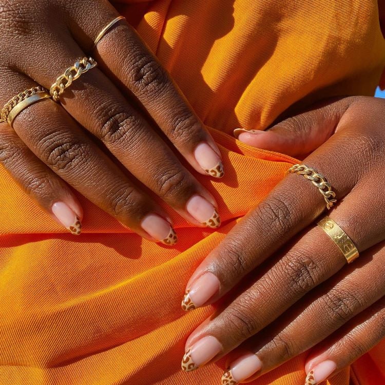 A imagem mostra duas mãos sobrepostas com unhas bem cuidadas, pintadas de marrom claro. As mãos estão adornadas com delicadas joias douradas e repousam sobre um tecido laranja vibrante, criando um contraste interessante com o tom de pele mais escuro das mãos.