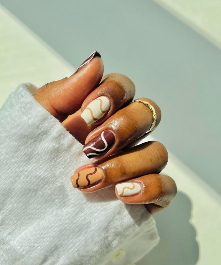 A imagem mostra uma mão com unhas pintadas artisticamente com esmalte marrom, branco e bege, formando desenhos abstratos. A luz solar destaca o brilho do esmalte e a textura da pele. Um anel dourado adorna o dedo anelar, complementando a elegância das unhas.