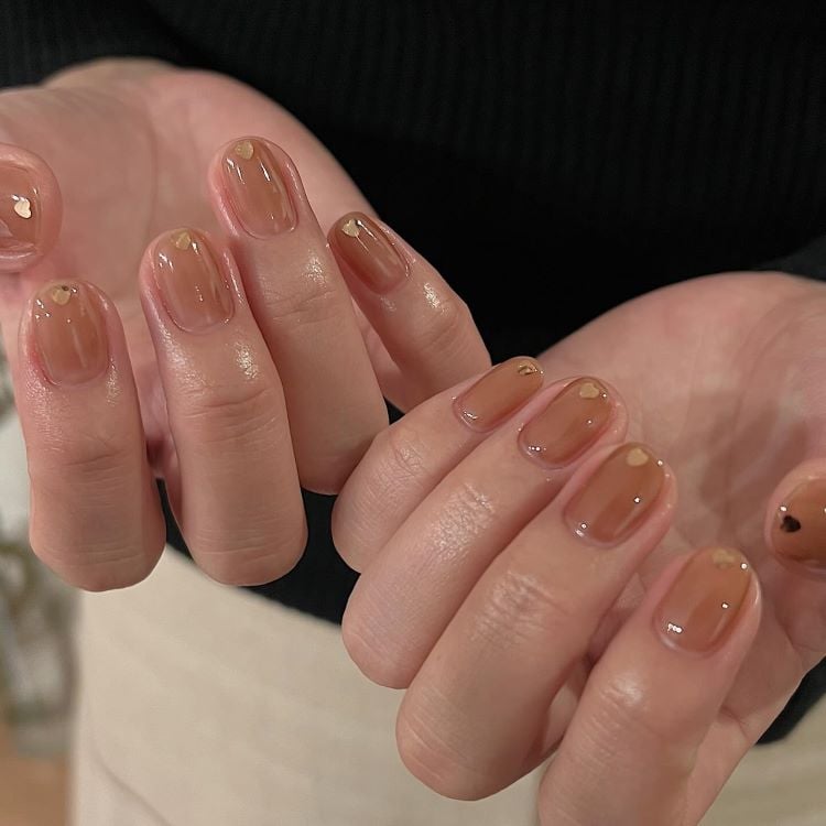 A imagem mostra um par de mãos com unhas pintadas em um tom de marrom claro, apresentando um acabamento brilhante. Algumas unhas possuem pequenos adornos dourados próximos às cutículas, e uma unha marrom apresenta um único strass. O fundo é desfocado, destacando as unhas.