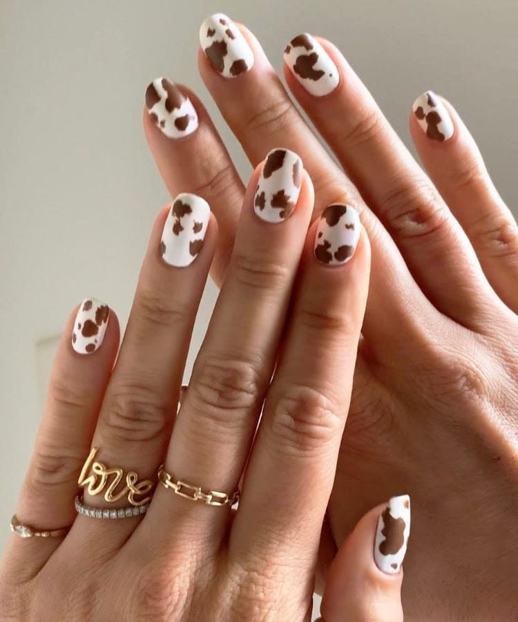 A imagem mostra um par de mãos com unhas pintadas em um estilo que imita a pelagem de uma vaca, combinando manchas marrons e brancas. As unhas estão bem cuidadas e o design é criativo e moderno. Uma das mãos exibe anéis dourados delicados, incluindo um com a inscrição “love”.