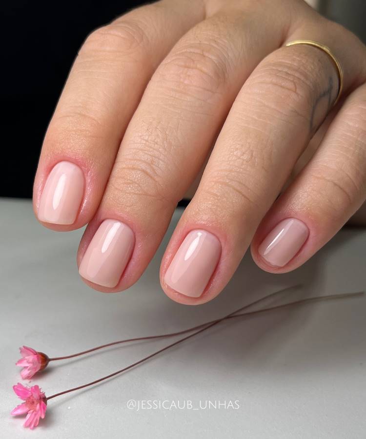 unhas curtas, nudes, elegantes. Mulher usa aliança dourada no dedo anelar. Sua mão está sob uma superfície com duas pequenas flores cor de rosa.