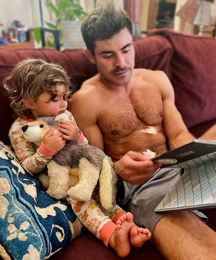 Ator Zac Efron sem camisa ao lado de criança, sentados em sofá, lendo um livro