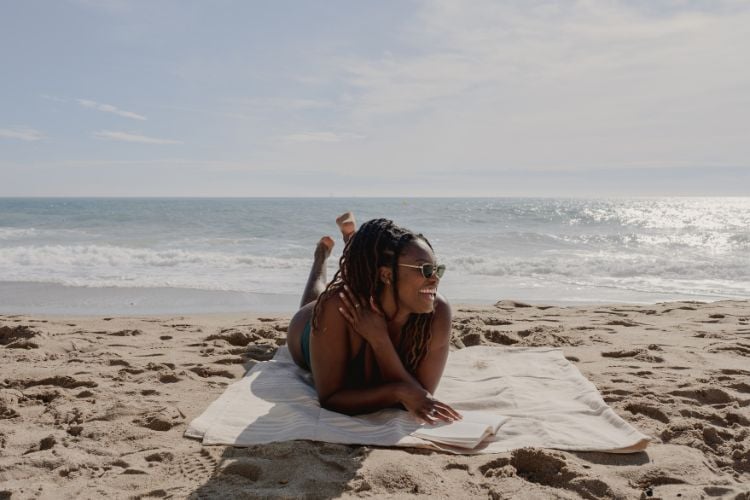 Mulher negra deitada em toalha de praia enquanto lê um livro