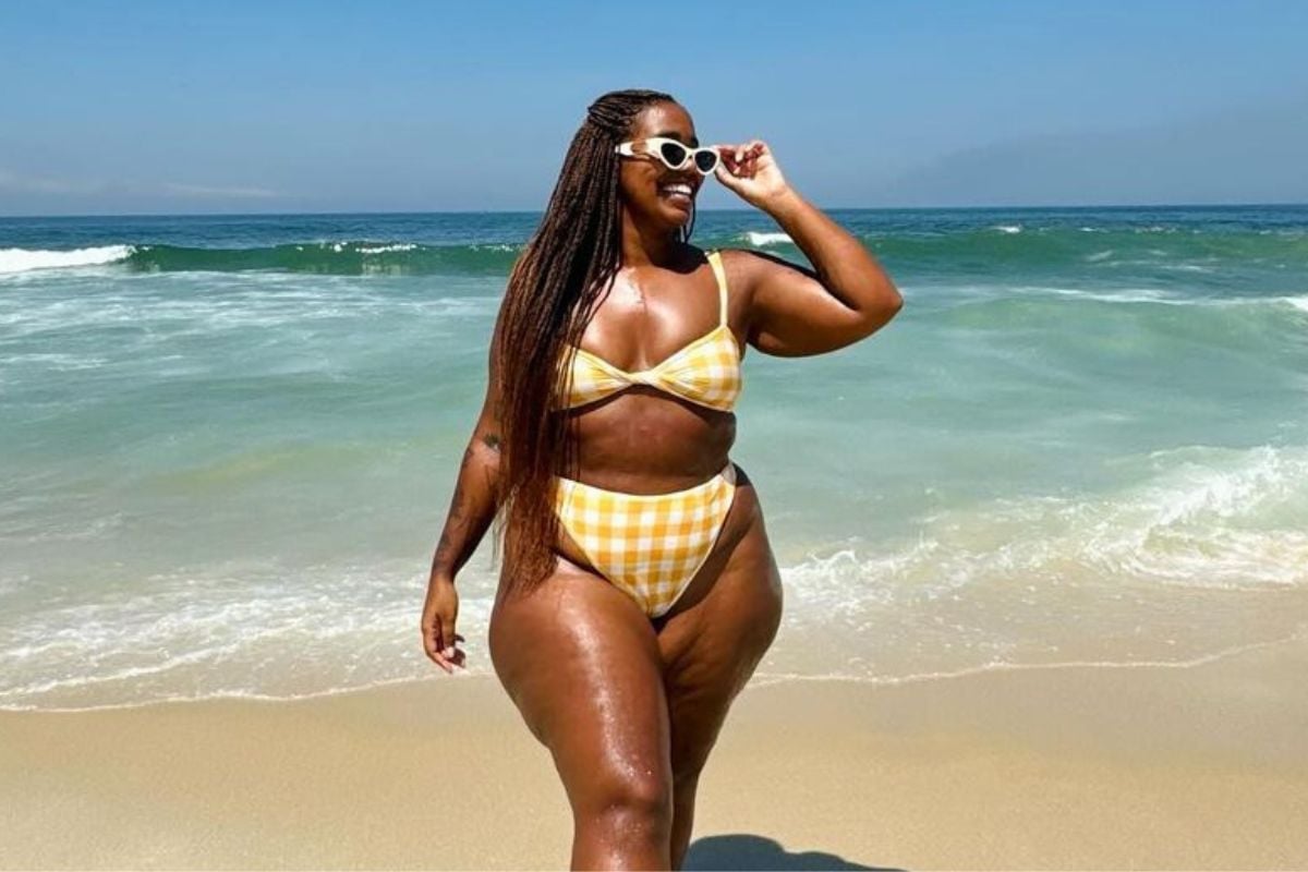 Foto da influencer negra plus size Mari Lobo na praia usando óculos de sol de armação branca e biquíni amarelo manteiga que está entre as tendências de moda praia para o verão 2025