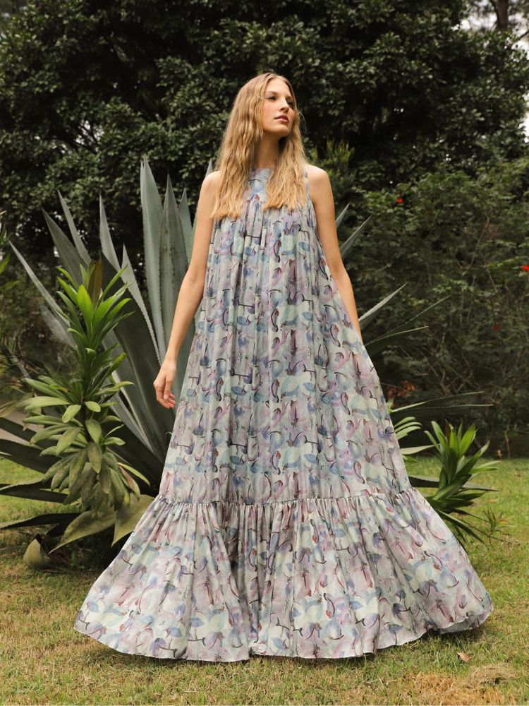 Mulher loira em pé em jardim usando vestido longo com estampa floral em tons de azul e roxo