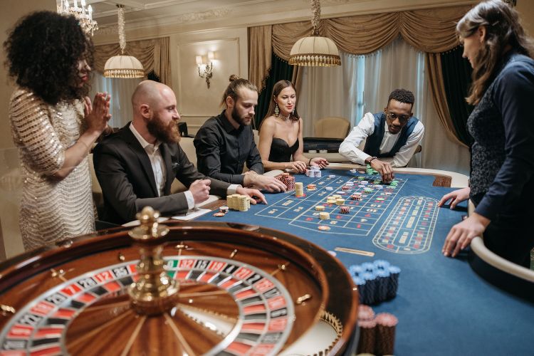 Foto de 5 pessoas jogando roleta usando roupas de gala em um cassino com uma dealer