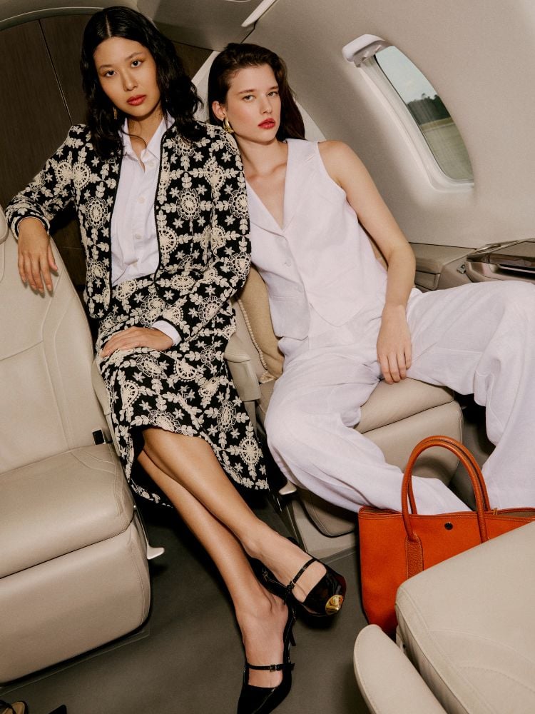 Duas modelos em um avião, uma usando conjunto de blazer e saia bordados, outra de colete e calça branca da coleção Jet Set, da Amissima Verão 2025