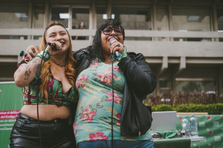 Foto de duas rappers latinas usando blusas floridas e sorrindo enquanto cantam abraçadas