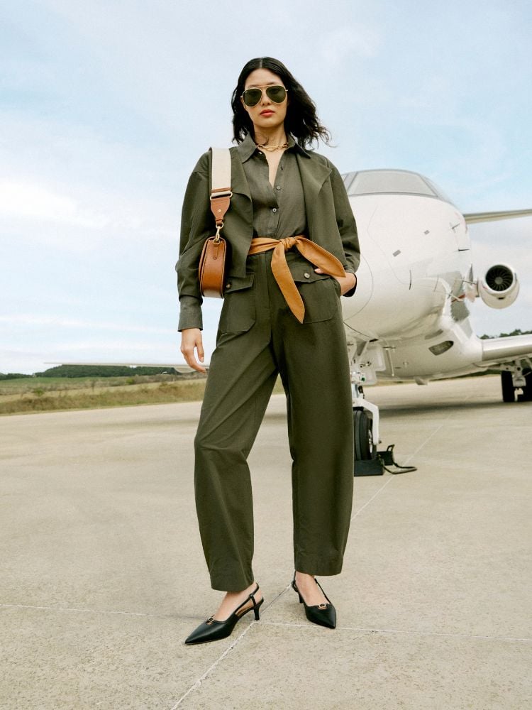 Modelo em aeroporto usando conjunto de blazer e calça verdes, com camisa da mesma cor e cinto de couro marrom