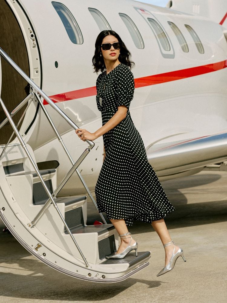 Mulher subindo escada de avião usando vestido preto de bolinhas brancas e sapato de salto prata