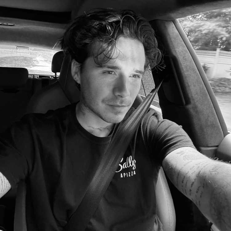 A imagem em preto e branco mostra Brooklyn Beckham dirigindo um carro. Ele tem cabelo ondulado e está usando uma camiseta com a inscrição "Sally Apizza". O cinto de segurança está afivelado, e ele parece estar olhando para fora, com uma expressão tranquila. O fundo mostra a parte interna do carro e uma vista desfocada da rua.