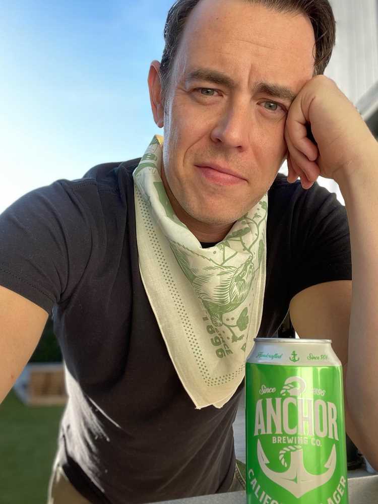 A imagem mostra um homem (Colin Hanks) com cabelo castanho escuro e curto, apoiando o rosto em uma das mãos. Ele veste uma camiseta preta e um lenço claro com detalhes verdes no pescoço. À sua frente, há uma lata verde de Anchor Brewing Co. O fundo é um céu azul claro, sugerindo que ele está ao ar livre.