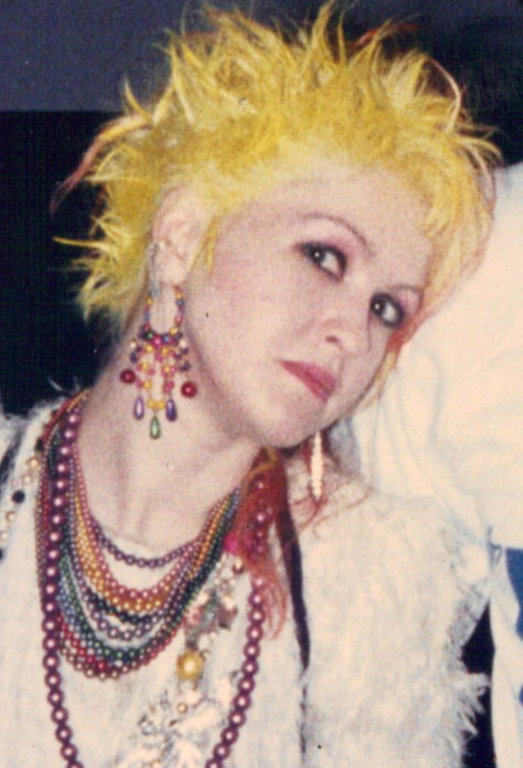 Cyndi Lauper, uma das famosas dos anos 80, exibe seu estilo excêntrico com cabelo amarelo espetado, maquiagem vibrante e acessórios coloridos. Ela usa várias colares e brincos chamativos, representando a moda ousada da época. A imagem captura sua personalidade única e a energia que a tornaram um ícone pop.