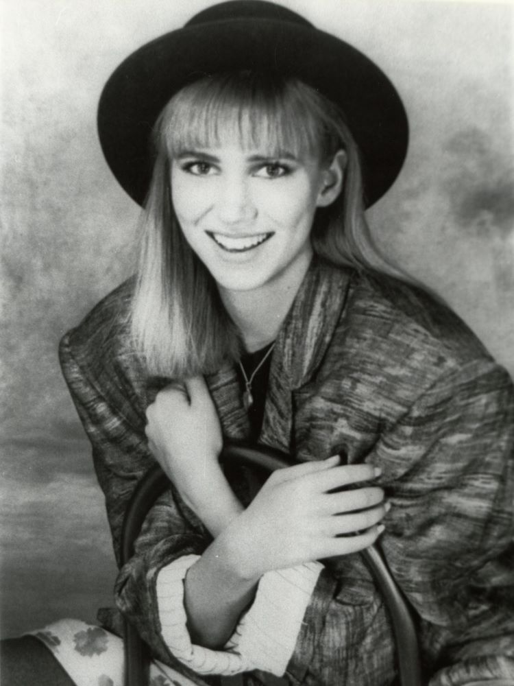 Debbie Gibson, uma das famosas dos anos 80, em uma foto promocional da época. Ela está sorrindo, usando um chapéu preto e uma jaqueta ampla, com um visual típico da moda juvenil dos anos 80. A imagem captura sua juventude e popularidade como cantora adolescente.