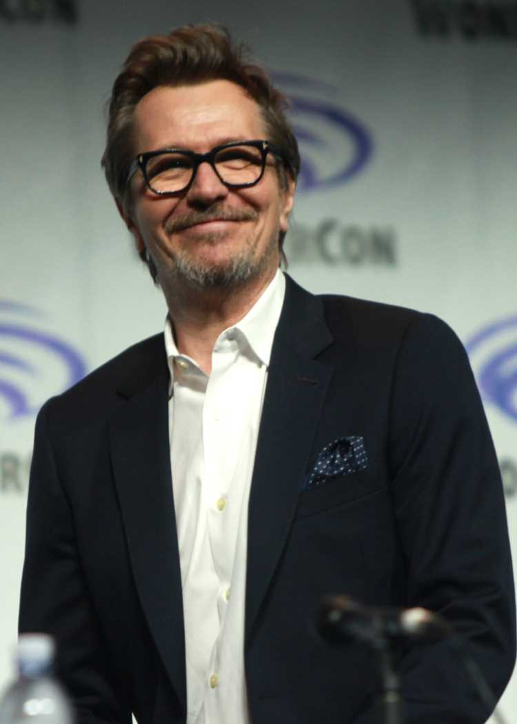 A imagem mostra Gary Oldman sorridente, usando óculos, com cabelo castanho claro e barba. Ele está vestindo um terno escuro com uma camisa branca e um lenço de bolso azul com bolinhas. O fundo parece ser de um evento, com um logotipo parcialmente visível.