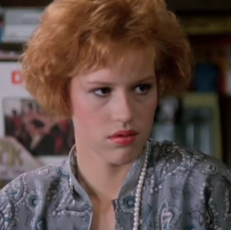 Molly Ringwald, uma das famosas dos anos 80, em uma cena do filme "A Garota de Rosa Shocking". Ela está com cabelo ruivo curto e expressão séria, usando uma blusa cinza estampada e colar de pérolas. A imagem captura seu papel icônico e a moda da época.