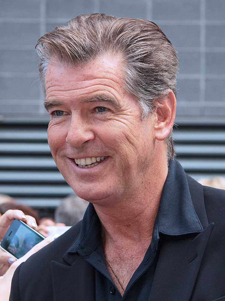 Na imagem, há um homem (Pierce Brosnan) sorrindo, com cabelo grisalho penteado para trás. Ele está vestindo uma camisa preta e um blazer escuro. O fundo é desfocado, com pessoas e celulares visíveis, sugerindo que ele está em um evento público ou ao ar livre.