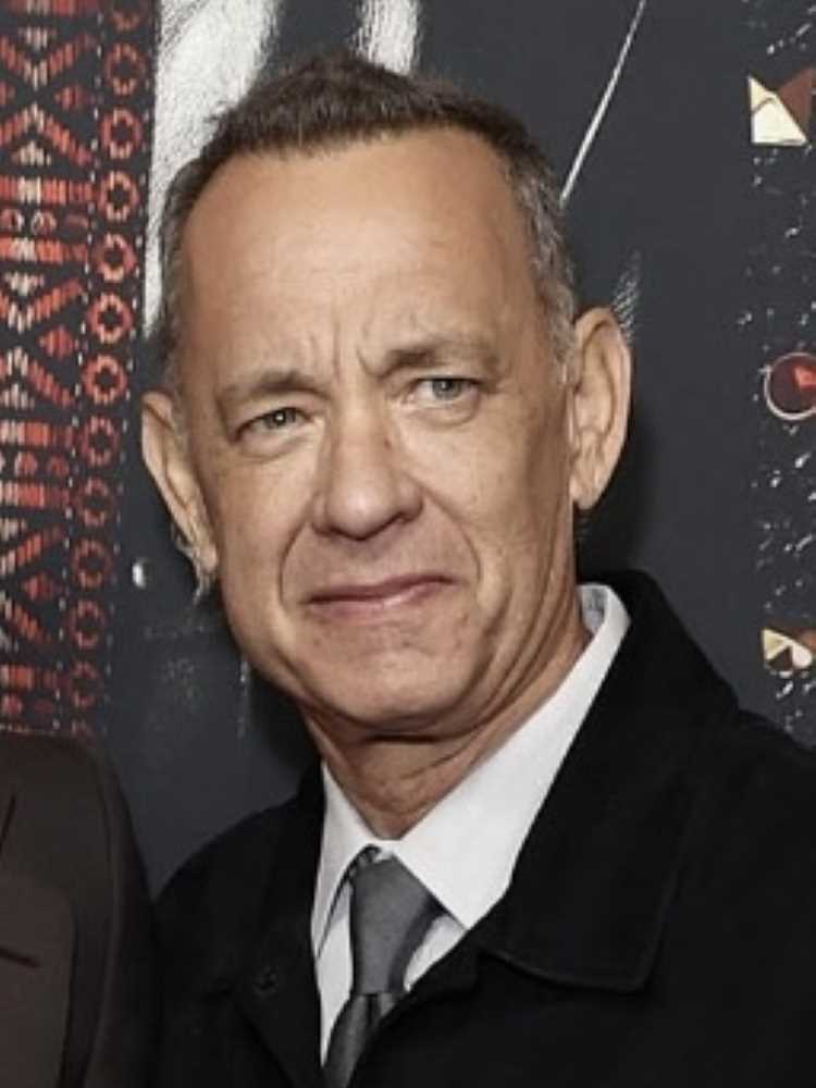 A imagem mostra um homem de meia-idade (Tom Hanks) com cabelo curto e grisalho. Ele está usando um terno escuro com uma camisa branca e gravata cinza. O fundo parece ser de um evento, com um padrão decorativo e cores escuras. Ele tem uma expressão neutra no rosto.