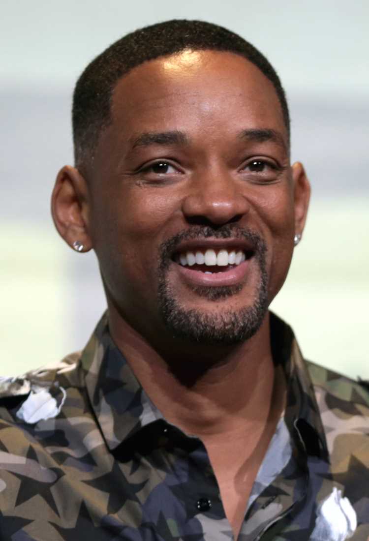 A imagem mostra Will Smith sorridente com cabelo curto e barba bem aparada. Ele está usando uma camisa com estampa camuflada em tons de verde, cinza e preto. Ele também está usando brincos pequenos em ambas as orelhas. O fundo é neutro e desfocado.