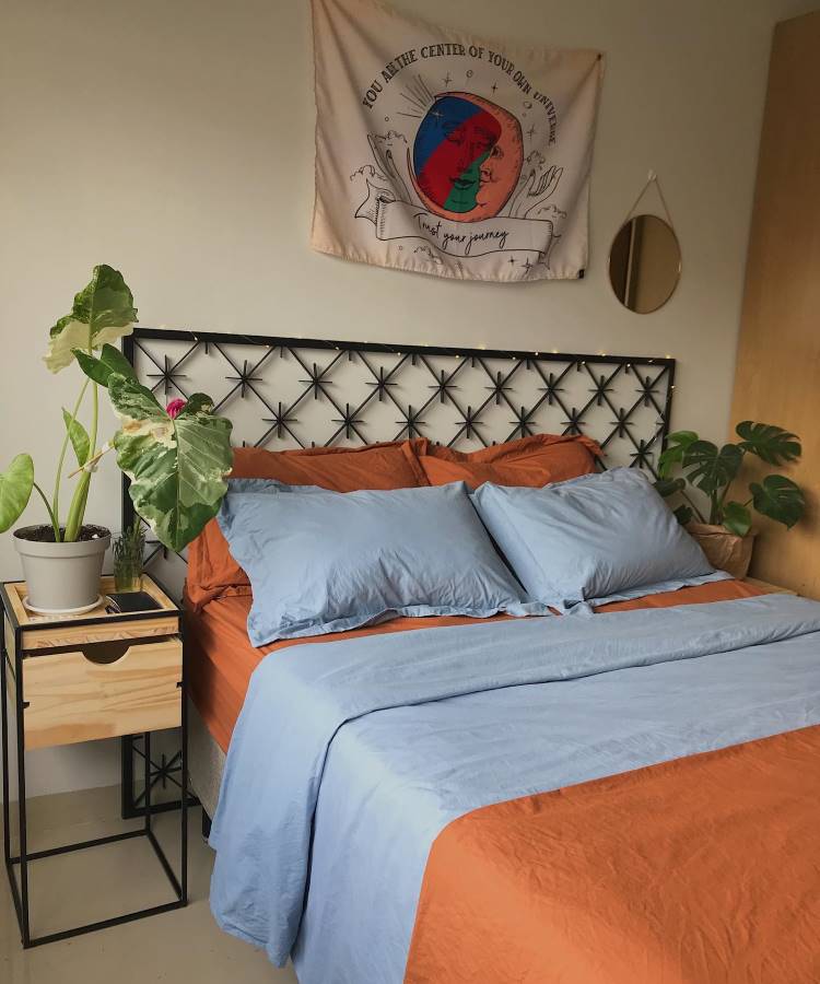 Um quarto com uma cama de lençóis azuis e cobertor laranja. Ao lado, uma alocasia em um vaso cinza está sobre um criado-mudo de madeira. As folhas são grandes e verdes, adicionando um toque natural ao ambiente. Uma tapeçaria colorida decora a parede acima da cama.